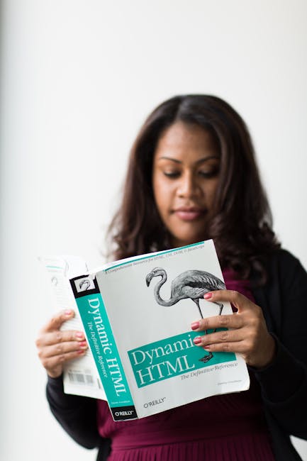 The image shows a woman with long, dark hair wearing a black blazer and a burgundy top, standing indoors against a plain white background. She is focused on reading a large book titled 'Dynamic HTML' 